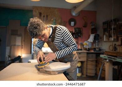 Sanding wood with orbital sander at workshop. Attentive man carpenter polishes wooden seat of a future chair with electric sander. Carpentry workshop. Furniture production - Powered by Shutterstock