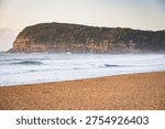 The sandstone cliffs of the headland of Second Point glow in morning light from Copacabana Beach on the Central Coast of New South Wales, Australia.