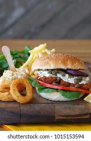 Sandwhich With Fried Chicken, Fried Onion Rings And Potatoes On A Wooden Board And Wooden Table, Close Up