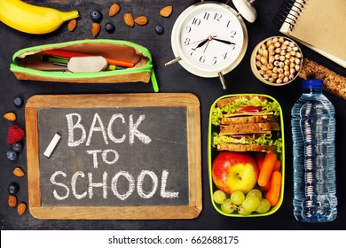Sandwich, apple, grape, carrot, berry in plastic lunch boxes, stationery and bottle of water on black chalkboard. Back to school concept. - Shutterstock ID 662688175