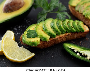 Sandwich With Avocado On A Dark Homemade Bread Made From Fresh Sliced Avocados On A Black Stone Background. Close-up. Slices Of Lemon, Cilantro And Jalapenos And A Half Avacado On The Background. 