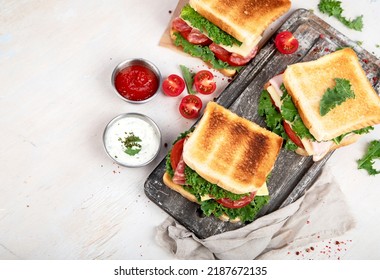 Sandwich With Cheese, Ham, Tomato And Fresh Salad, Made Of Whole Wheat Bread With A Crispy Crust On A Wooden Board With Sauces On A White Background, Top View With Copy Space.