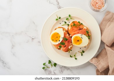 Sandwich with delicious toppings, smoked salmon, eggs, herbs and microgreens radish, black sesame seeds over white plate on white marble table background. Healthy open sandwich superfood. Top view.