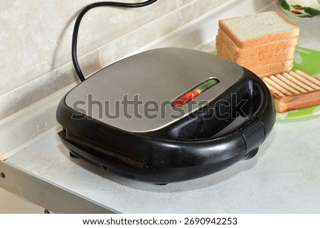 The sandwich maker toasts bread, the light on the top panel is on.