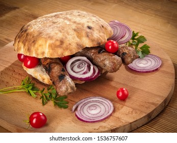 Sandwich with roasted bread bun, grilled cevapi, sliced red onion and cherry tomatoes on wooden board