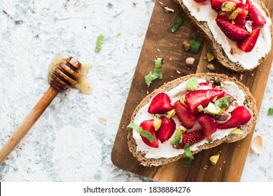 Sandwich with strawberries, soft cheese, pistachios, mint and honey on wooden board on grey background. Top view.
