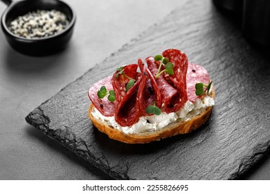 Sandwich, toast with sliced salami, pepperoni, cream cheese, sausage, microgreen on dark slate cutting board. Snack, bruschetta. Copy space, text
