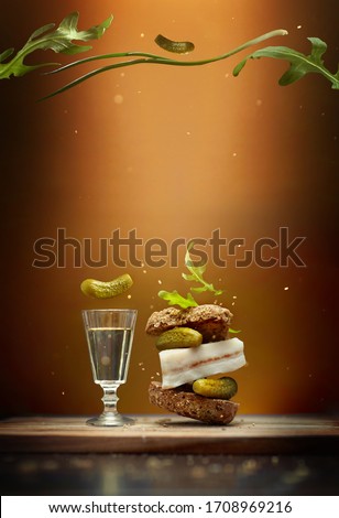 A sandwich in Ukrainian style with slice of bacon on brown background with flying ingredients pickles, spring onion and arugula  with a glass of vodka, ornamented with greenery on the top