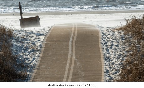 Sandy beach access mat with wind-blown sand leading to choppy ocean waves on a sunny, blustery day, bordered by dry dune grass, depicting a powerful natural environment.