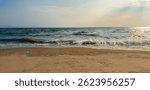 A sandy beach with gentle ocean waves lapping against the shore under golden afternoon sunlight, while faint distant hills line the horizon beneath a lightly clouded sky. 