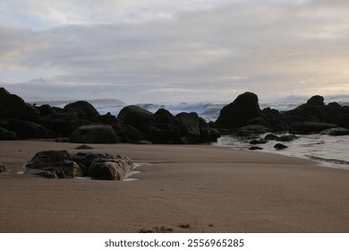 A sandy beach with large, dark rocks scattered along the shore. Waves are crashing against the rocks under an overcast sky, with the horizon visible in the background.

 - Powered by Shutterstock