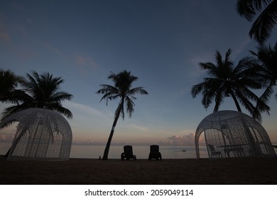 A Sandy Beach With Palm Trees And Two Lounge Chairs In The Evening