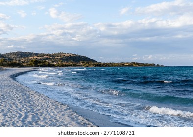 Sandy beach at S'Ena e Sa Chitta, Sardinia. Calm waves along the coastline. Clear blue sea under a cloudy sky. Rocky landscape in the distance.