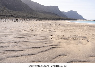 Sandy beach showcasing wind-swept patterns, a calm turquoise ocean, and a dramatic chain of volcanic mountains under a clear sky