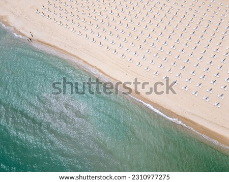 A sandy beach stretches along the seaside, adorned with empty sun loungers. Aerial view