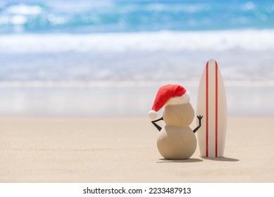 Sandy Christmas Snowman is watching the waves, standing on beautiful beach with a surf board - horizontal version - Powered by Shutterstock