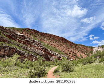 A sandy hiking trail winds through green shrubs beneath a red-and-white layered sandstone canyon slope under a partly cloudy sky.