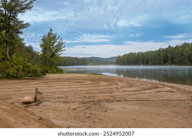 A sandy lakeside beach with surrounding trees. Lake is calm and stretches toward a distant tree-covered horizon, with a partly cloudy sky. Fog rises from the water. A rope is in the foreground. - Powered by Shutterstock