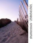 A sandy path filled with footprints leading to Horseneck Beach in Westport Massachusetts. The path is lined by a leaning fence. Sand dunes are on the other side of the path. It is early morning.