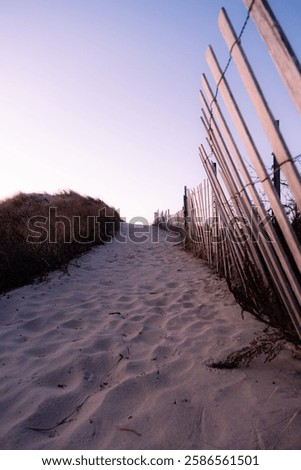 A sandy path filled with footprints leading to Horseneck Beach in Westport Massachusetts. The path is lined by a leaning fence. Sand dunes are on the other side of the path. It is early morning.