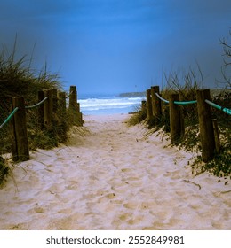 A sandy pathway flanked by wooden posts and green ropes, leading to a beach with crashing waves under an overcast sky. - Powered by Shutterstock