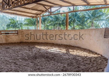 A sandy round pen or paddock with a rustic, covered structure featuring masonry walls and wooden beams. 