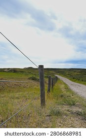 A sandy rural path bordered by a wooden fence and grassy terrain under a partly cloudy sky, creating a calm open‑landscape mood.