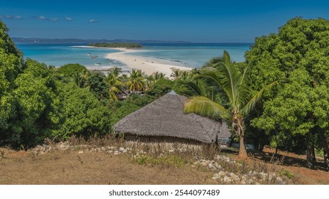 A sandy winding spit leads to a small green island in the aquamarine ocean. Tiny silhouettes of people on the beach. Boats on the water. Lush vegetation, thatched roof of a hut on the foreground - Powered by Shutterstock