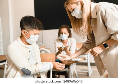 Sanitary measures against Covid19 coronavirus spread at school. Teacher using sanitizer with her students pupils schoolchildren before lesson class wearing protective face masks. - Powered by Shutterstock