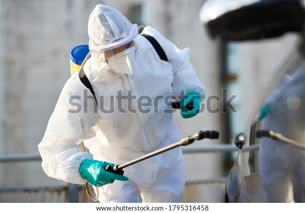 Sanitation worker in hazmat suit spraying\
car with disinfectant due to COVID 19 epidemic.\
