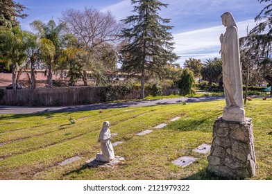 Santa Barbara, California, USA - February 8, 2022: Calvary Cemetery. Our Lady of Lourdes statue group on green burial lawn with trees and housing in back under blue cloudscape.