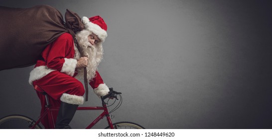 Santa Claus carrying a heavy sack with gifts for Christmas and riding a bicycle, banner with blank copy space