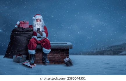 Santa Claus delivering gifts on Christmas Eve, he is sitting on a chimney and connecting online with his smartphone - Powered by Shutterstock