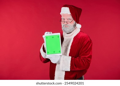 Santa Claus dressed in traditional red suit holding a tablet with a green screen while standing against a red background - Powered by Shutterstock