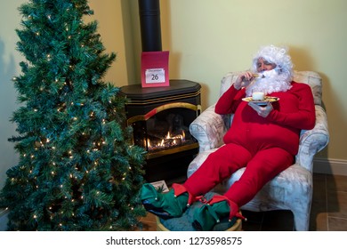 Santa Claus Eating Cookies And Milk In House Wearing Red Long Johns Relaxing The Day After Christmas.