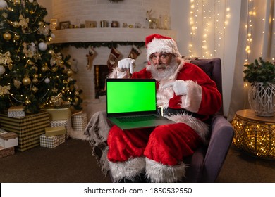 Santa Claus in glasses sitting in his rocker near christmas tree. holds a laptop with green screen and attracts attention to it. Christmas spirit, holidays and celebrations concept 4k footage - Powered by Shutterstock