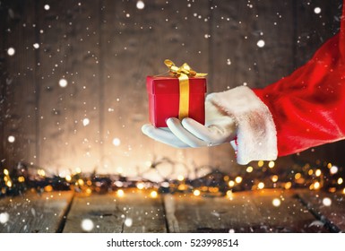 Santa Claus Hand Holding Christmas Gift Box Over Wooden Background. Proposing Product. Advertisement Gesture Presenting Point. Decorated Christmas Tree With Winter Holiday Gifts. 