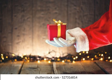 Santa Claus hand holding Christmas Gift box over wooden background. Proposing product. Advertisement gesture presenting point. Decorated Christmas tree with Winter Holiday Gifts