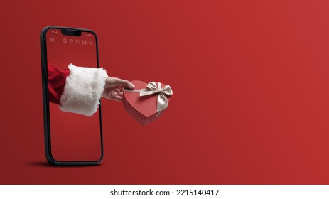 Santa Claus hand holding a heart shaped Christmas gift and coming out from a smartphone screen, blank copy space - Powered by Shutterstock