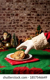 Santa Claus' Hand Reaching For A Home Made Cookie, Christmas Table Setting. Brick Wall In The Background, Xmas Ornaments Decorating. Vertical, Text Space.