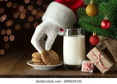 Santa Claus hand takes chocolate chip cookie. Snacks and glass of milk for Santa under Christmas tree. Golden lights in background. Copy space, - Powered by Shutterstock