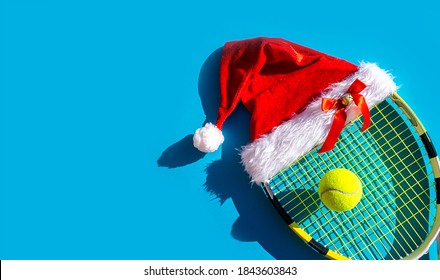 Santa Claus hat on tennis racket and ball on blue background. Merry Christmas and Happy New Year tennis concept. Close up, sport lifestyle, funny. 