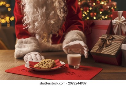 Santa Claus having a delicious snack, he is eating cookies and drinking milk, Christmas and holidays concept - Powered by Shutterstock