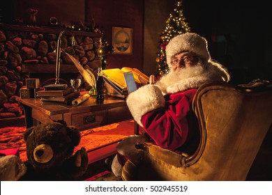 Santa Claus in his work shop using a cell phone while doing his naughty or nice list and giving a thumbs up to the camera