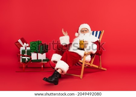 Santa claus holding glass of beer on deck chair near sleigh with presents on red background