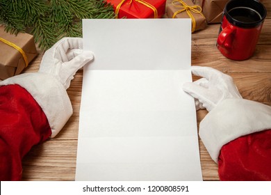 Santa Claus Holding Letter On Wooden Table With Gift Boxes And Christmas Tree And Cup Of Hot Coffee Or Tea.  Mockup Blank