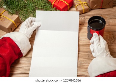 Santa Claus Holding Letter On Wooden Table With Gift Boxes And Christmas Tree And Cup Of Hot Coffee Or Tea.  Mockup Blank