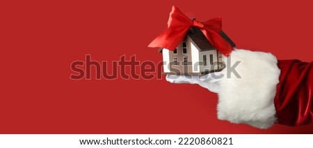 Santa Claus holding model of house with ribbon bow on red background with space for text. Concept of real estate