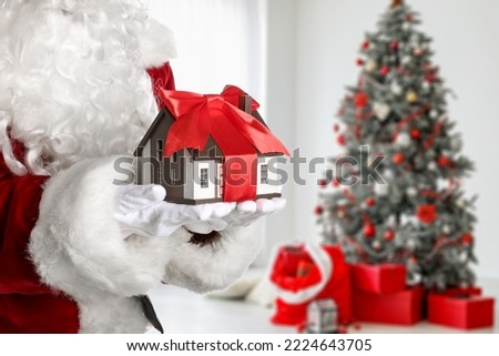 Santa Claus holding model of house in room decorated for Christmas. Concept of real estate