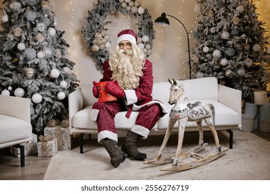 Santa claus holding red christmas gift box sitting on sofa with rocking horse near decorated christmas tree - Powered by Shutterstock